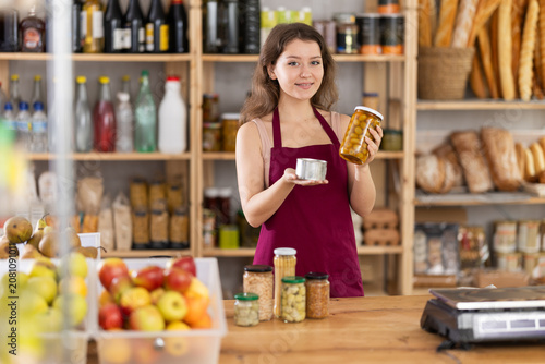 Positive young saleswoman is holding canned products standing at the counter in market
