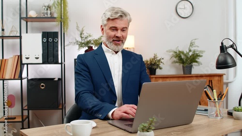 Middle-aged businessman at home office scrolls laptop news feed sees sharp sales rise lifts thumbs up celebrating. Freelancer guy at table plans next goals analyzing graphs forecasting profit growth.