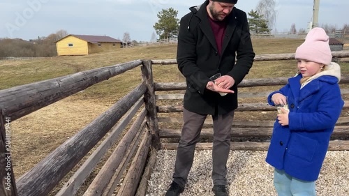 Happy father and little daughter feeding a goat with snacks from cups at a rural petting zoo farm.