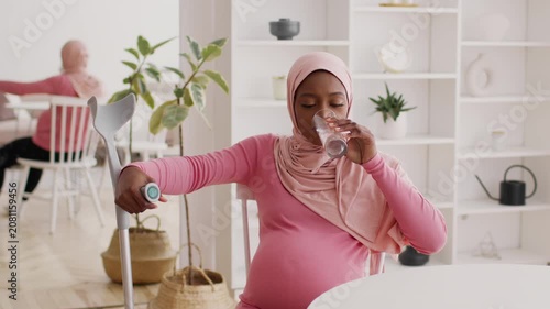 Focused Pregnant Black Woman Using Crutch And Drinking Water At Home, Sitting At Kitchen Table In Modern Environment, Prioritizing Health And Wellness During Pregnancy.