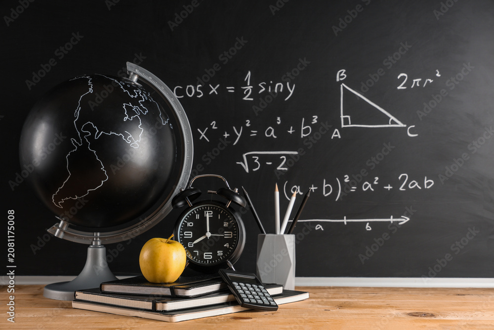 Obraz premium Apple, globe, alarm clock and stationery on desk near chalkboard with formulas. Back to school concept