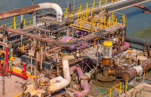 High-angle view of complex industrial marine machinery with weathered, rusty pipes, pumps, and yellow handrails on a vessel or terminal deck near the water during the daytime