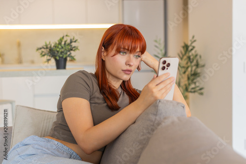 Fotografie Young woman at home browses news on smartphone too long feels bored annoyed and drained