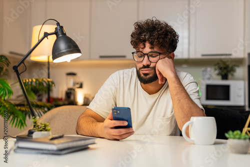 Obraz na płótnie Arabian man at home scrolls smartphone news feels bored stressed, negative expression, sighs wants rest