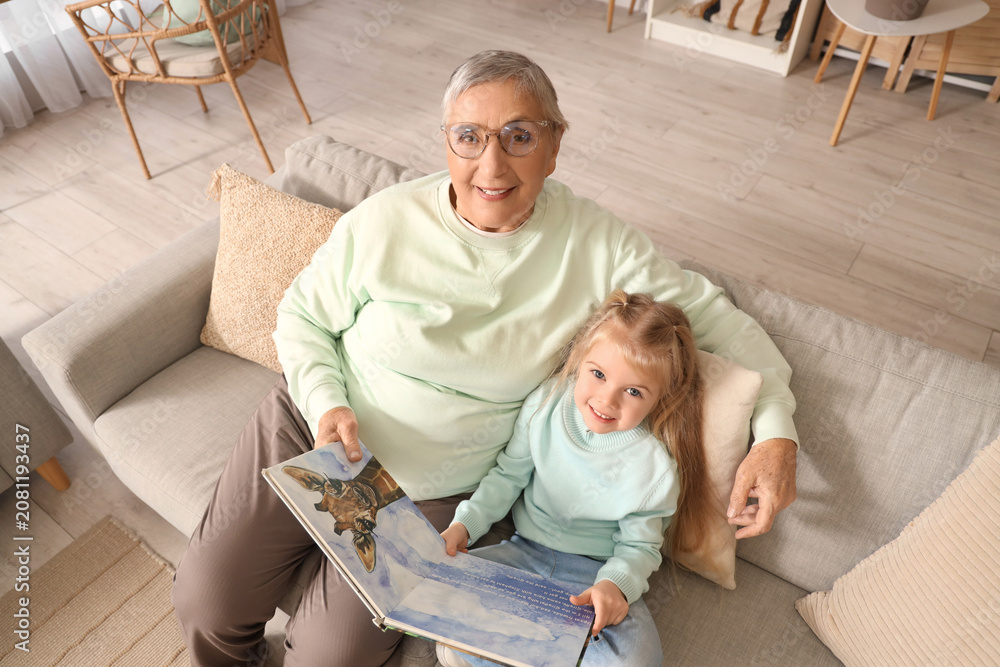 Obraz premium Cute little girl with her granny reading story on sofa at home