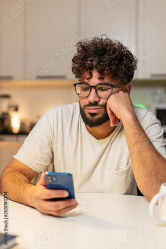 Bild auf Leinwand Arabian man at home scrolls smartphone news feels bored stressed, negative expression, sighs wants rest