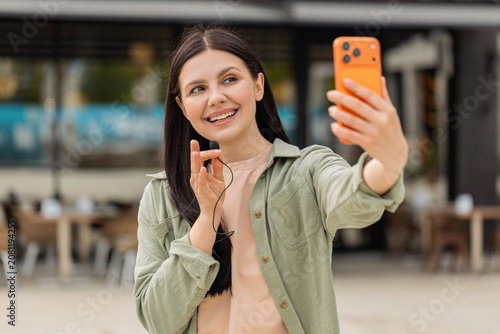 Young woman filming location review walking in city street with smartphone and lavalier microphone. Girl tourist speaking confidently outdoors showing cafe background for travel blog and urban vlog.