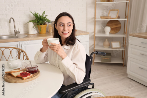 Happy young woman in wheelchair drinking tea at home