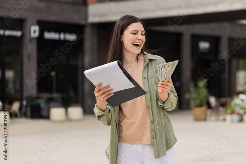 Young woman celebrating successful deal walking in city street after agreement and payment success. Girl tourist smiling outdoors showing joy with cash and documents after profitable contract result.
