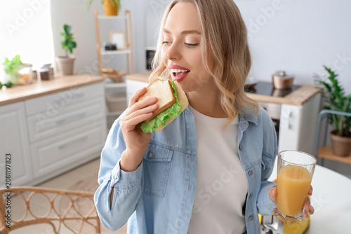 Young beautiful woman with glass of orange juice eating tasty sandwich in kit...