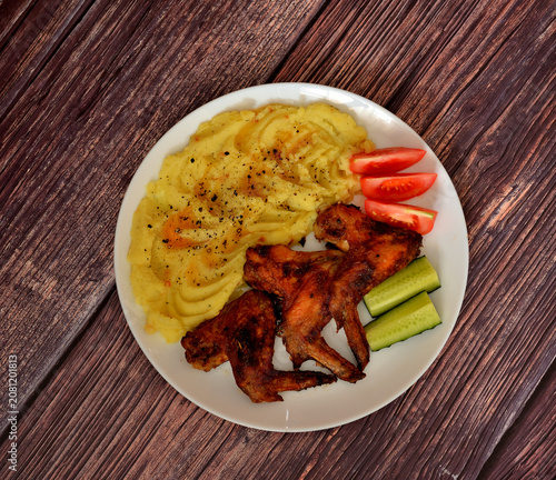 A plate of delicious lunch, baked chicken wings with mashed potatoes, tomatoes and fresh cucumber on a wooden table.
