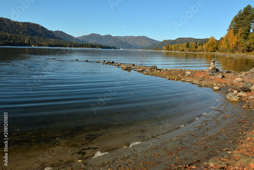 A rocky spit with a stone pyramid extends from the shore to the middle of a mountain lake on a sunny autumn morning.