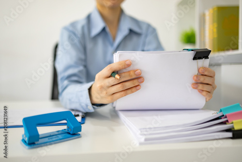 Professional woman organizes documents with colorful clips. She holds a marker, ready to annotate papers. Stacked sheets and vibrant clips add visual interest. Focused demeanor suggests productivity
