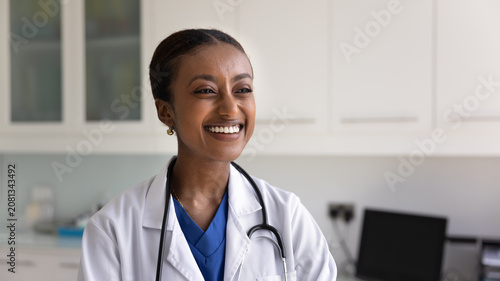 Foto Happy beautiful African American doctor woman in lab coat and stethoscope posing in medical office, looking away with toothy smile