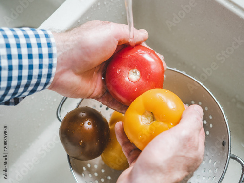 Male hands wash ripe tomatoes under running water over a metal colander in a kitchen sink. Concept of healthy eating and using natural sweeteners. Ideal for nutrition, vegan diet, and cooking content