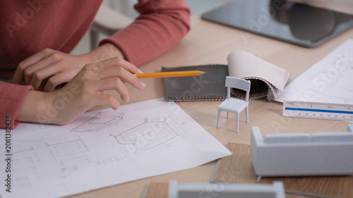 A designer's hand holding a pencil reaches toward a detailed furniture sketch while a miniature chair and fabric swatches sit on the desk.
