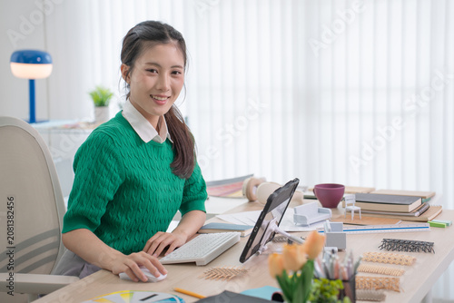 A cheerful interior designer sits at her desk using a keyboard and mouse surrounded by fabric samples and small furniture models.