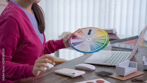 A designer's hand extends a color wheel reference chart beside an open laptop while a miniature armchair sits on the desk.