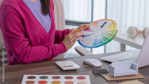 A close up of a designer's hand holding a pencil and rotating a colorful color wheel chart at her desk with a laptop nearby.