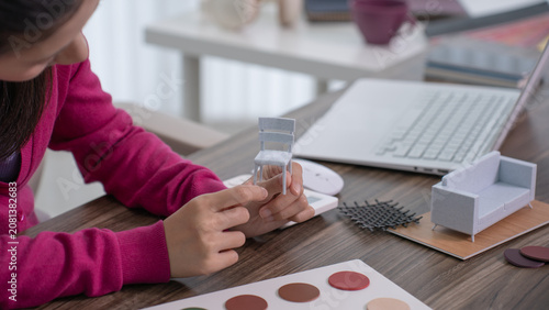 A smiling designer touches a color palette board while holding a miniature chair model in her other hand at a cluttered desk.
