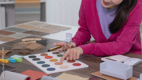 A designer's hand extends a color wheel reference chart beside an open laptop while a miniature armchair sits on the desk.
