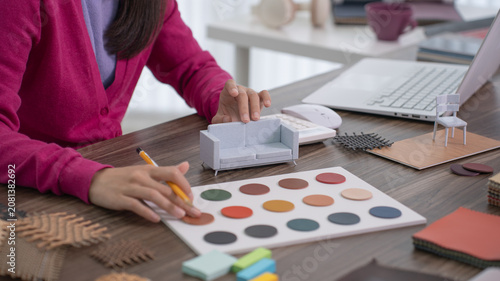 A close up of a designer's hand examining a small scale white chair model near a laptop and color palette board.