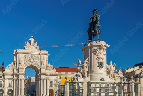 A prominent bronze equestrian statue on a decorative stone pedestal standing alongside a grand historic triumphal arch under a clear blue sky in Lisbon
