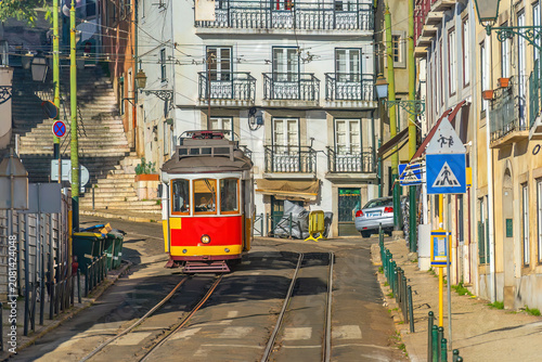 A traditional red vintage tram ascends a steep, narrow cobblestone street lined with historic buildings in Lisbon