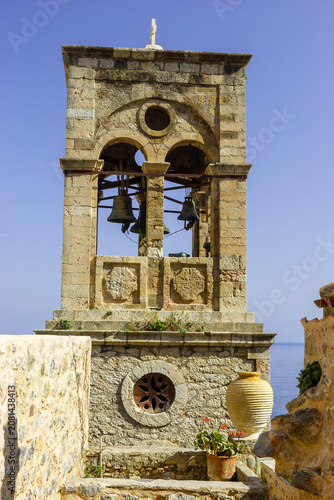 Fotografía A stone bell tower with bells and a cross on top stands against a clear blue sky, with the sea visible in the background