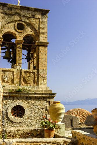 Fotografía A bell tower with bells and a large amphora stand in front of a tiled dome and the sea in Monemvasia, Greece
