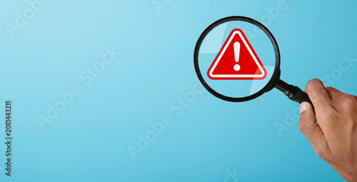 Tableau sur toile Hand holding a magnifying glass over a red warning icon, illustrating the proces