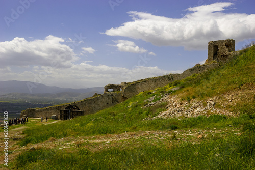 A group of tourists explore the ancient ruins of a stone castle on a grassy hillside under a blue sky with white clouds.