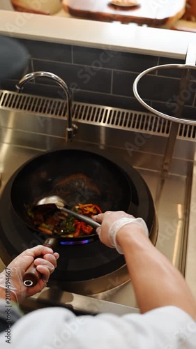 Restaurant cook with a metal ladle stir-frying fresh vegetables in a wok over a gas  burner. Chef prepares asian food in a restaurant.