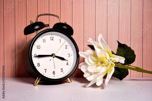 Black analog alarm clock with plastic flower decoration isolated on horizontal wooden wall and white table background.