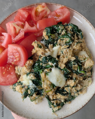 Close-up of a healthy breakfast plate featuring fluffy scrambled eggs mixed with spinach, melted mozzarella cheese, and fresh tomato slices