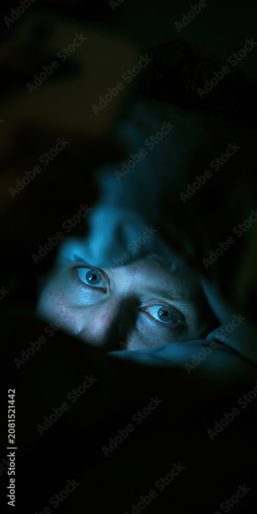 Obraz premium Person lying in bed with blue smartphone light on face in dark bedroom, digital insomnia concept, copy space