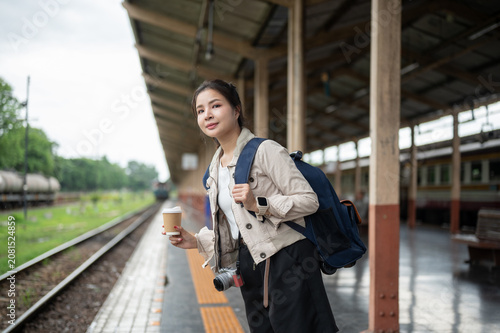 Asian woman traveler carrying a backpack waiting with coffee beside railway tracks at train station.