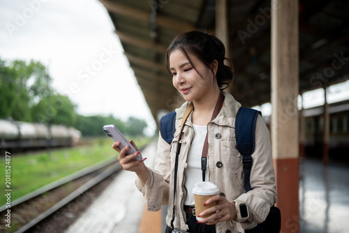 Asian woman traveler checking smartphone while holding coffee at railway station during a waiting.