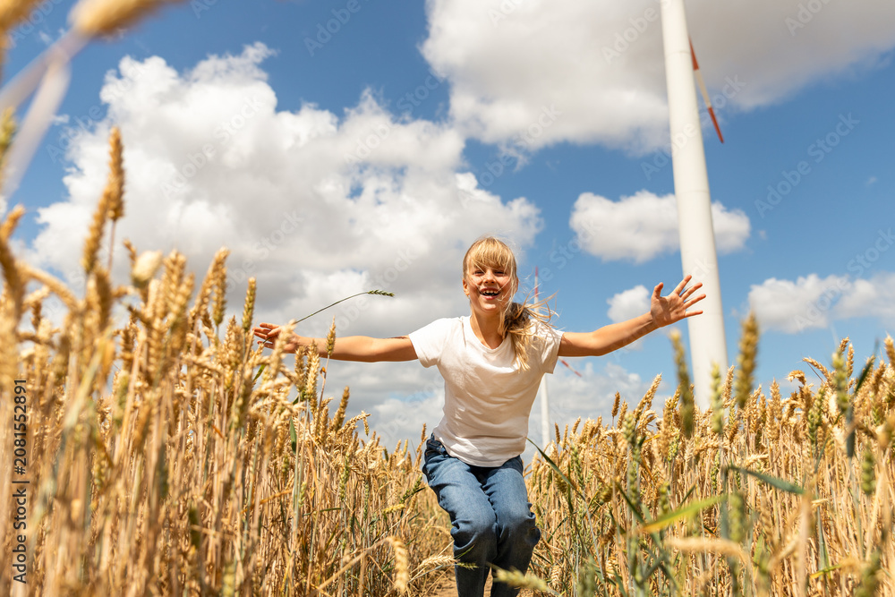 Fototapeta premium Laughing girl runs through golden wheat field with open arms, wind turbine rising behind under blue sky. Sunny scene suggests childhood joy, clean energy, harvest, nature, freedom, sustainability