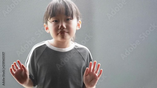Little girl stands near window screen. Hands raised, eyes calm, expression neutral. Soft light illuminates her quiet pose. Ideal for family, children, or quiet moments concept