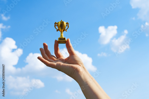Hand holds and raised small cup trophy, holding cup against clean sky backgrund. Consolation prize award and victory concept.