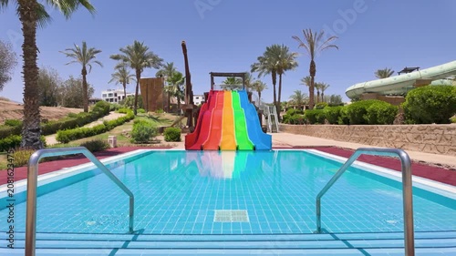 Slow motion footage of a colourful rainbow water slides beside a swimming pool in Sharm El Sheikh Egypt surrounded by palm trees and tropical resort gardens beneath a bright blue sky