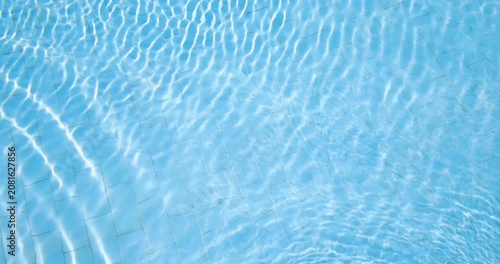 Slow motion footage of crystal clear blue swimming pool water gentle ripples reflecting bright sunlight across tiled surfaces creating a relaxing summer holiday background with shimmering patterns