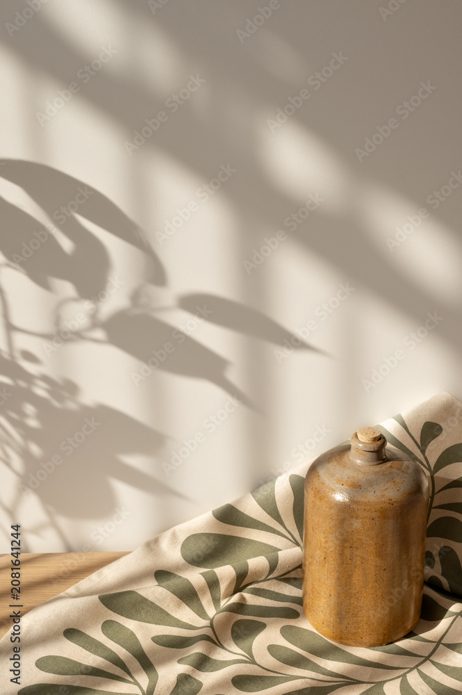 Fototapeta premium Crafted clay bottle, linen napkin on table, soft natural sunlight shadows on plain beige wall background. Aesthetic home interior decor in Mediterranean or rustic style