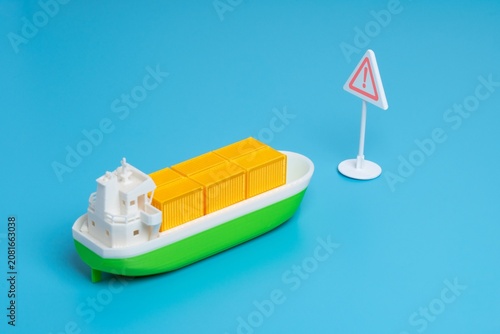 Miniature cargo ship and a triangular warning sign. Maritime hazards, supply chain risks, shipping alerts, trade sanctions concept.