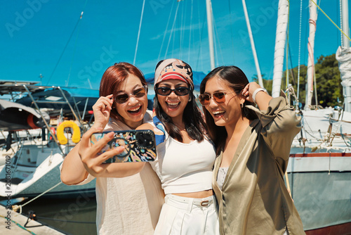 Three happy young Asian women taking a selfie together at a marina with luxury yachts in the background.