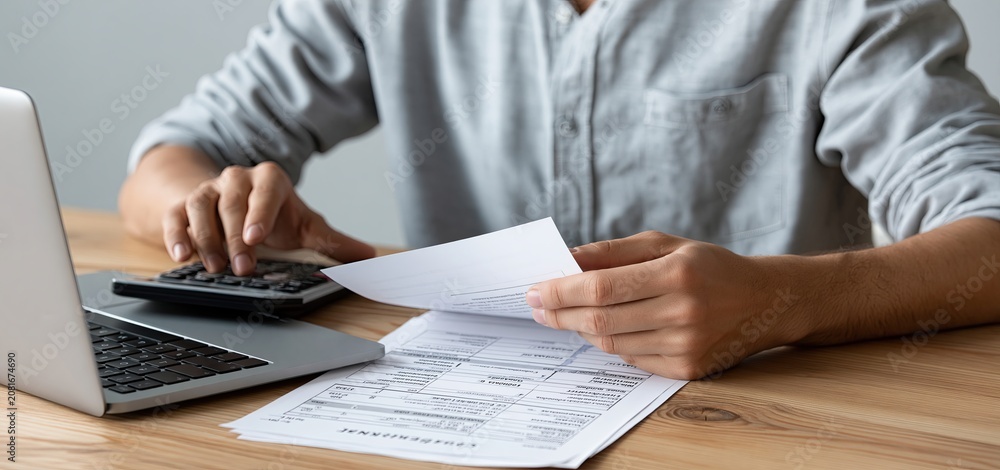 Fototapeta premium Person calculating expenses with financial documents, laptop, and calculator on a wooden office desk.