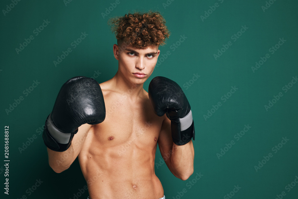 Obraz premium Young male boxer with curly hair wearing black boxing gloves in fighting stance against green background. Athletic shirtless man training for boxing or martial arts.