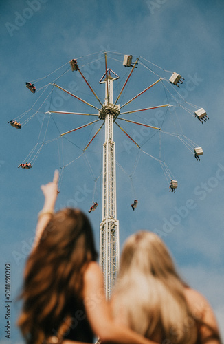 Фотография Two young women gaze up at a thrilling carnival ride against blue sky