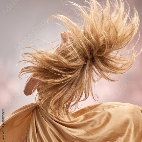 Joyful young woman in golden baroque satin dress with long flowing blonde hair tossed dramatically mid-air, smiling against soft studio background, perfect for hair care beauty advertising